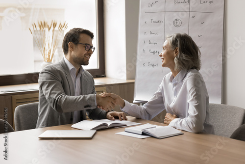 Happy mature businesswoman giving handshake to younger colleagues man, discussing agreement at meeting table. Employer and candidate shaking hands after successful job interview