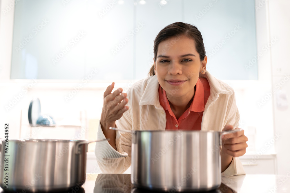 Young woman chef standing near stove and cooking.Happy woman looking and smelling tasting fresh delicious from soup in a pot with steam at white interior kitchen Young woman chef standing near stove and cooking.Happy woman looking and smelling tasting fresh delicious from soup in a pot with steam at white interior kitchen