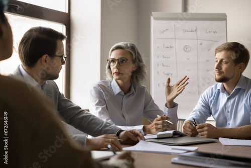 Serious elder business leader woman discussing project, brainstorming on strategy with team at table. Mature corporate coach giving seminar to office staff, talking to colleagues in boardroom. photo