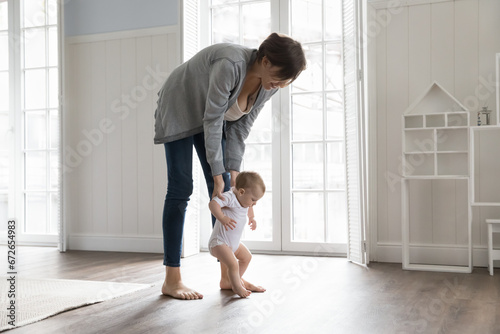 Cute toddler in bodysuit holding loving mothers hands and makes first steps in cozy bedroom, walk on warm floor with underfloor heating system. Adoption, care, happy motherhood, growth and development