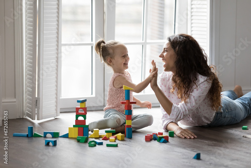 Happy mother playing wooden blocks with daughter, finish build tower, praising kid, giving high five settled down together on warm floor in living room. Playtime, help, happy motherhood, leisure games