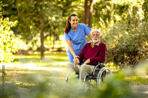 Female nursing assistant and elderly man in wheelchair enjoying autumn in the park and looking to the camera. photo