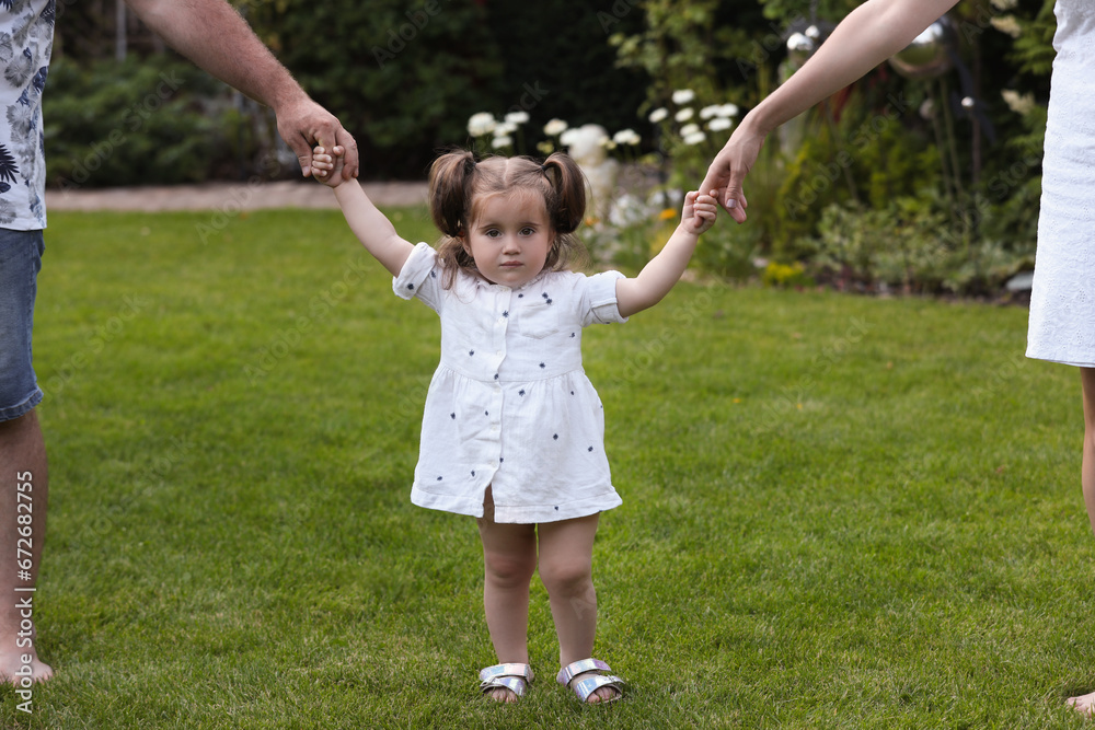 Parents supporting baby girl while she learning to walk on green grass in park Parents supporting baby girl while she learning to walk on green grass in park
