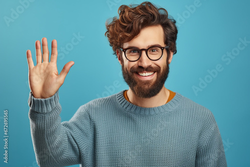 Young handsome man with beard wearing casual sweater and glasses over blue background Waiving saying hello happy and smiling, friendly welcome gesture
