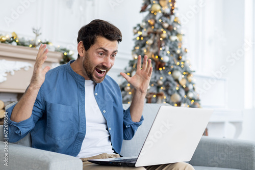 Man joyfully shouting at laptop screen got online win and victory, achievement results are good, celebrating triumph, sitting on sofa in living room, celebrating new year and christmas photo