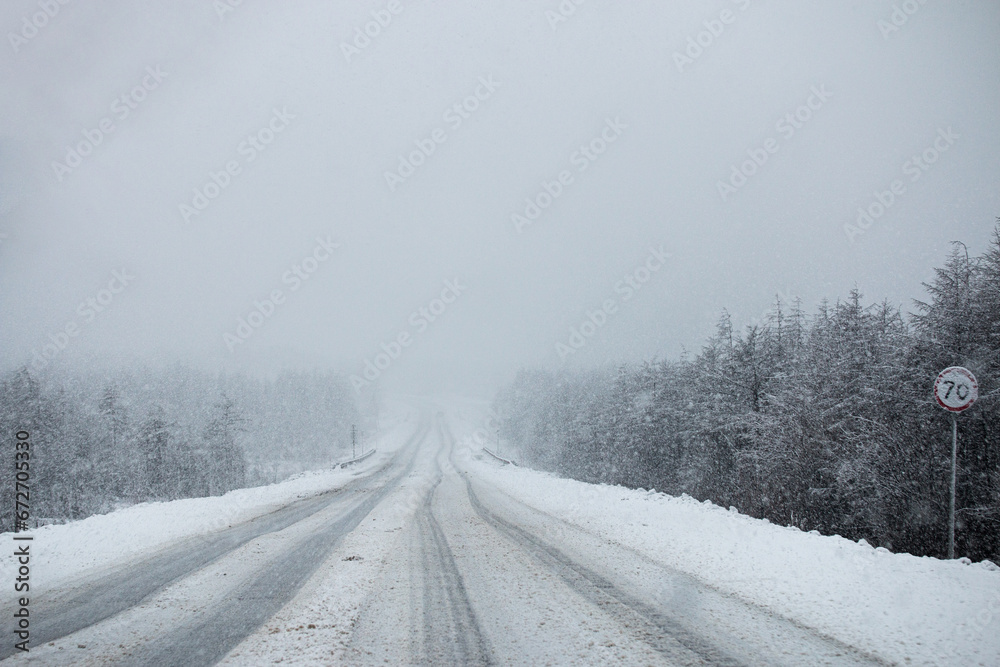 Obraz premium Photo of the winter road during the snowfall in Magadan, Russia. Snow showers on the trees and hills. Slush on the way. Fog and haze, low visibility due to snowstorm. Extreme weather conditions