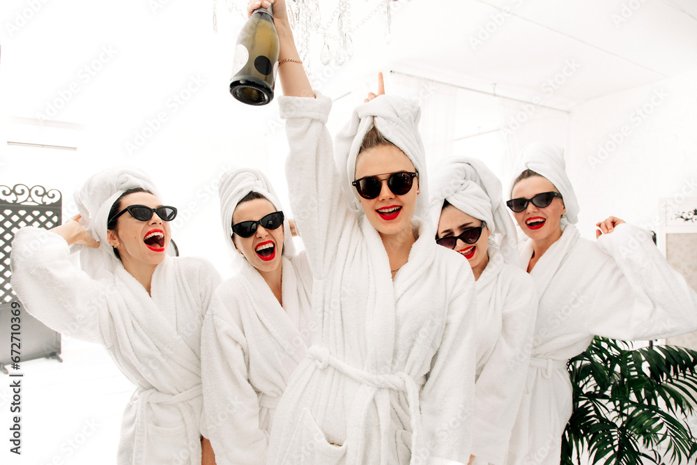 Cheerful cute multiracial young girlfriends in bathrobes having fun, holding glasses of champagne, sitting on bed in hotel. Concept for women's relaxation, spa and entertainment. - obrazy, fototapety, plakaty 