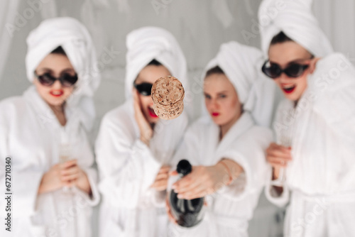 Photo with champagne opening and a flying cork. Women in bathrobes and glasses enjoy their vacation. Concept of entertainment and spa treatments for women.