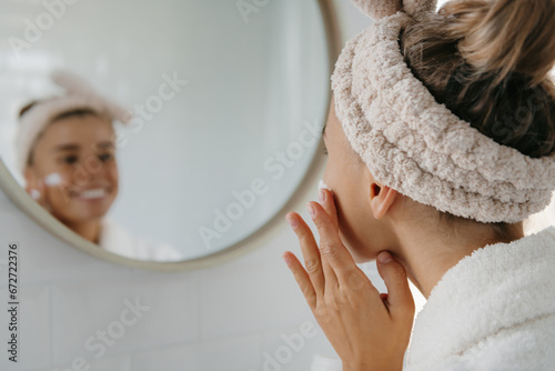 Beautiful young woman in bathrobe applying face cream while looking at her reflection in the mirror