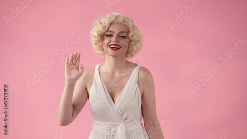 A woman dressed as waves her hand in greeting. A woman in a white dress and white wig with red lipstick on her lips in the studio on a pink background. photo