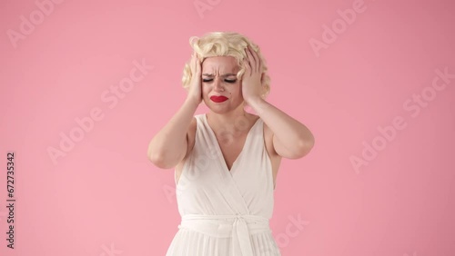Unhappy frustrated woman massaging her temples while suffering from a severe headache. Woman in image of in wig, white dress and with red lipstick on lips isolated on pink. photo