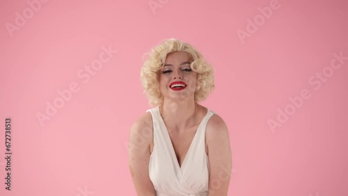 A woman in wig, white dress and with red lipstick on lips on pink background. The woman poses, portrays actress , copying her movements, holds the hem of her dress blown up by the wind.