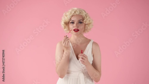 Young woman applies red lipstick on lips with makeup brush, and sends air kiss. Woman looking like in studio on pink background. photo