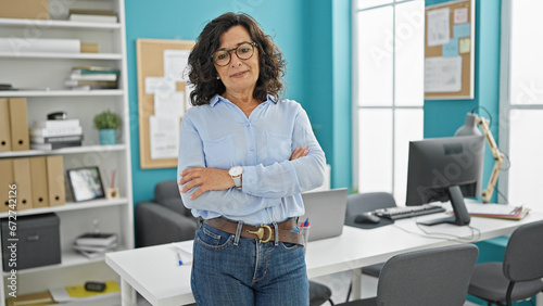 Middle age hispanic woman business worker standing with serious face and arms crossed gesture at the office