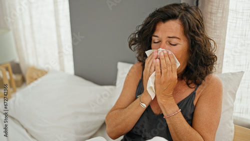 Middle age hispanic woman sitting on bed being sick sneezing at bedroom