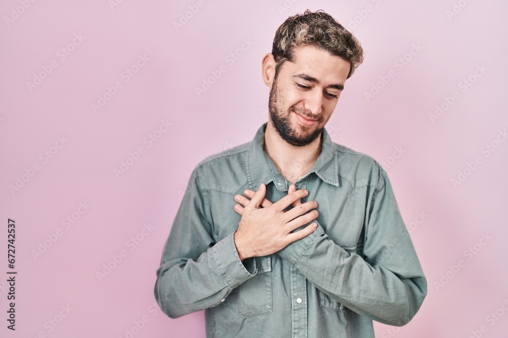 Hispanic man with beard standing over pink background smiling with hands on chest with closed eyes and grateful gesture on face. health concept. Hispanic man with beard standing over pink background smiling with hands on chest with closed eyes and grateful gesture on face. health concept.
