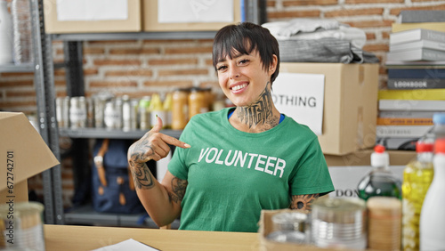 Young beautiful hispanic woman sitting on table pointing to volunteer uniform smiling at charity center