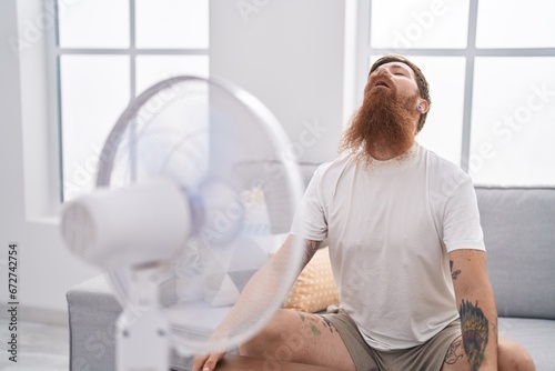 Young redhead man sitting on sofa using fan at home photo