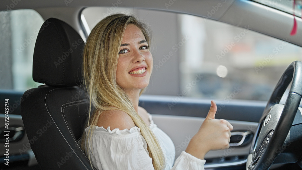 Young blonde woman driving car doing thumb up gesture at street