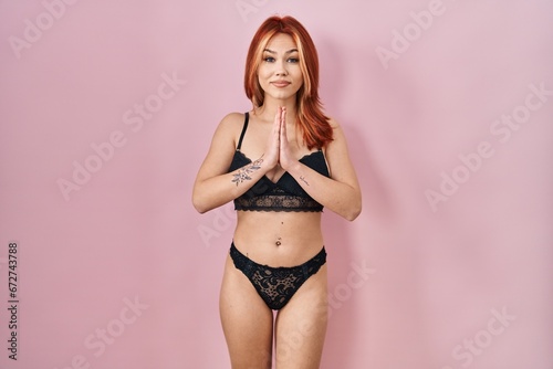 Young caucasian woman wearing lingerie over pink background praying with hands together asking for forgiveness smiling confident.