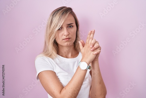 Young blonde woman standing over pink background holding symbolic gun with hand gesture, playing killing shooting weapons, angry face © Krakenimages.com