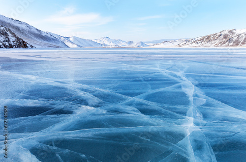 Scenic winter landscape of frozen Baikal Lake on cold sunny February day. Shallow bay is covered with blue ice with cracks against backdrop of snowy coastal mountains. Beautiful natural background