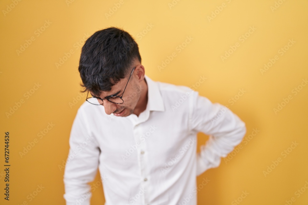 Young hispanic man standing over yellow background suffering of backache, touching back with hand, muscular pain Young hispanic man standing over yellow background suffering of backache, touching back with hand, muscular pain