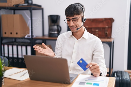 Young hispanic man working using computer laptop holding credit card pointing aside with hands open palms showing copy space, presenting advertisement smiling excited happy