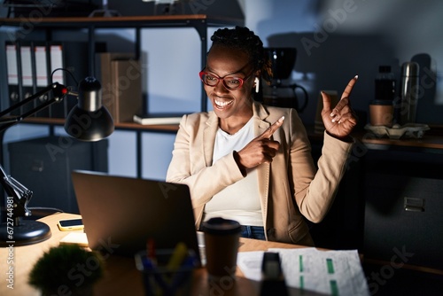 Beautiful black woman working at the office at night smiling and looking at the camera pointing with two hands and fingers to the side.
