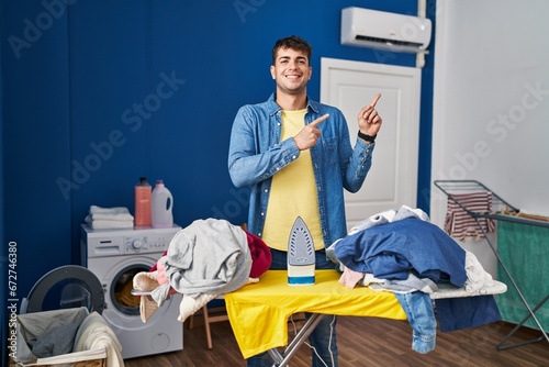 Young hispanic man ironing clothes at home smiling and looking at the camera pointing with two hands and fingers to the side.
