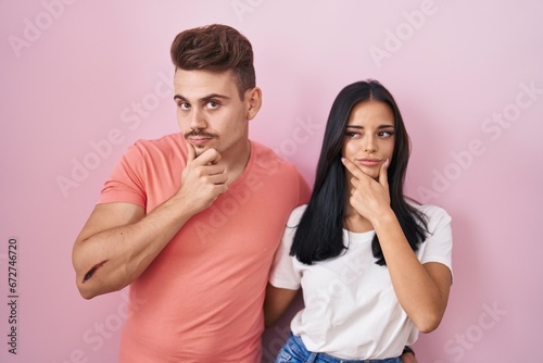 Young hispanic couple standing over pink background looking confident at the camera smiling with crossed arms and hand raised on chin. thinking positive.