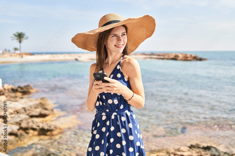 Young blonde woman tourist wearing summer hat using smartphone at beach