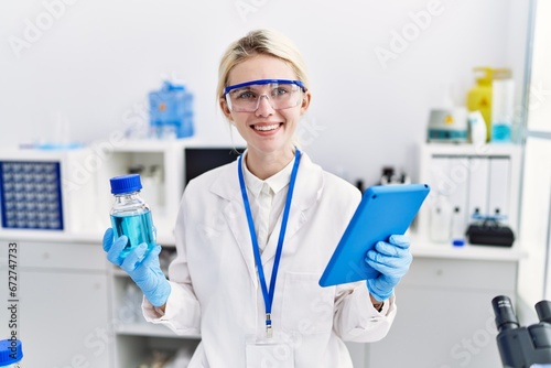 Young blonde woman scientist using touchpad holding bottle with liquid at laboratory