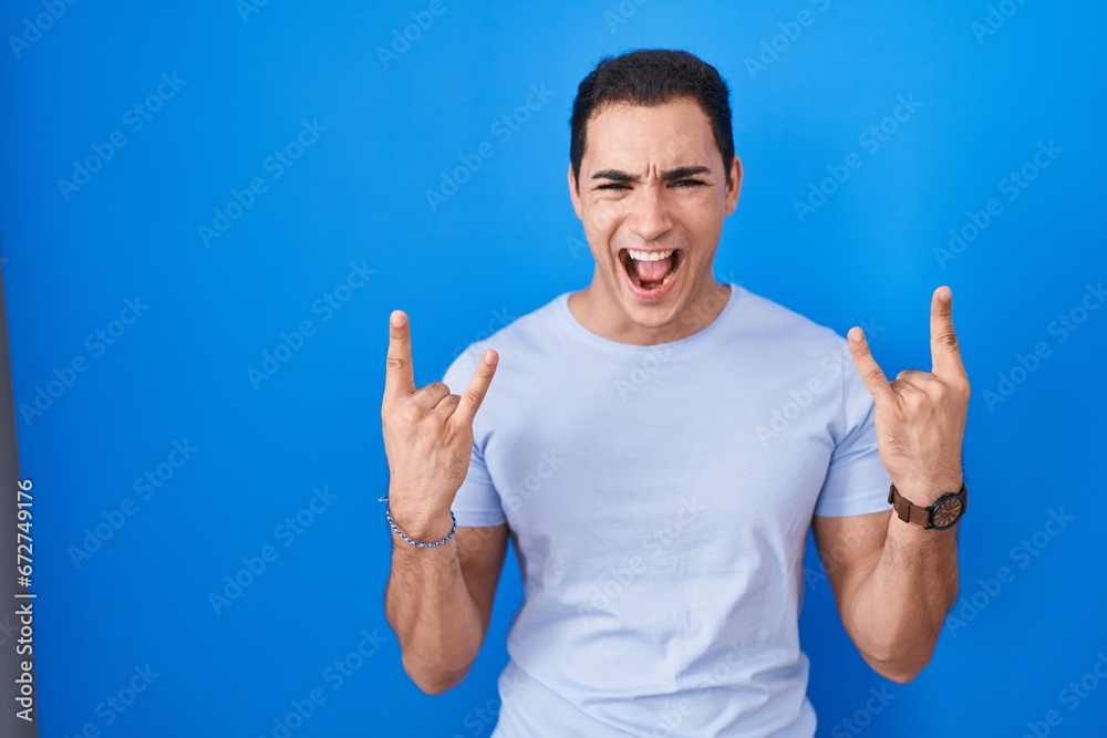 Young hispanic man standing over blue background shouting with crazy expression doing rock symbol with hands up. music star. heavy music concept. Young hispanic man standing over blue background shouting with crazy expression doing rock symbol with hands up. music star. heavy music concept.