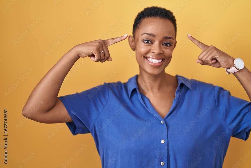 African american woman standing over yellow background smiling pointing to head with both hands finger, great idea or thought, good memory African american woman standing over yellow background smiling pointing to head with both hands finger, great idea or thought, good memory