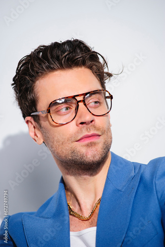 Fototapeta Naklejka Na Ścianę i Meble -  Trendy man with hairstyle posing in eyeglasses, blue jacket and golden chain on grey