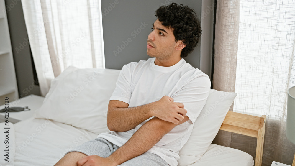 Young latin man scratching arm for itchy sitting on bed at bedroom Young latin man scratching arm for itchy sitting on bed at bedroom