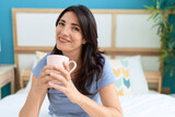 Middle age hispanic woman drinking cup of coffee sitting on bed at bedroom