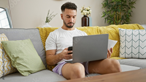 Young arab man using smartphone and laptop holding paper at home