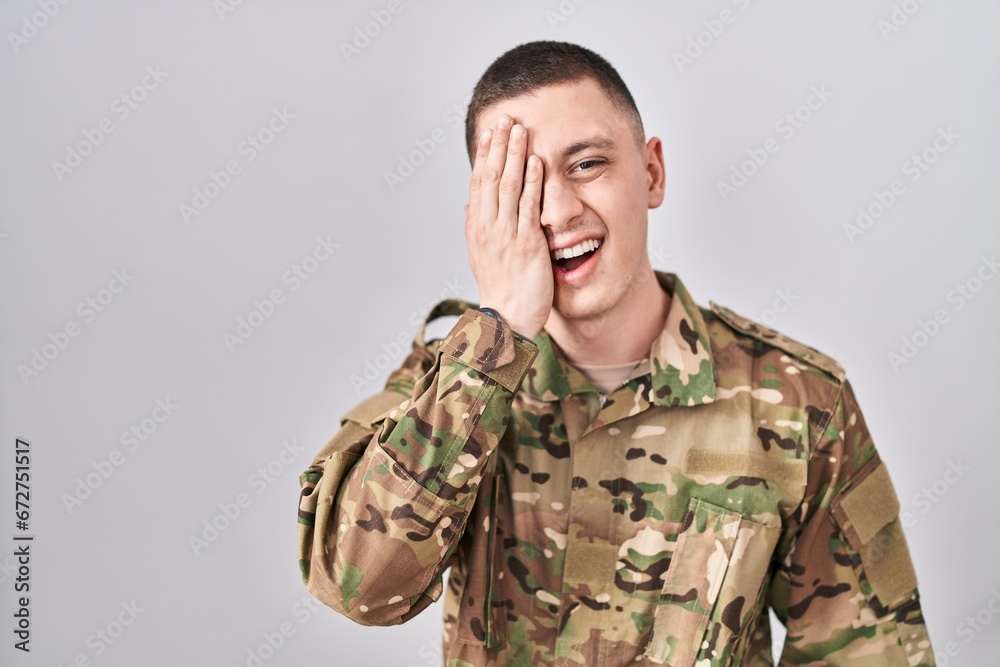 Young man wearing camouflage army uniform covering one eye with hand, confident smile on face and surprise emotion.