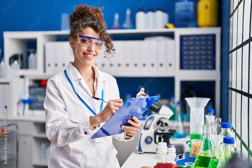 Young hispanic woman scientist smiling confident writing on document at laboratory