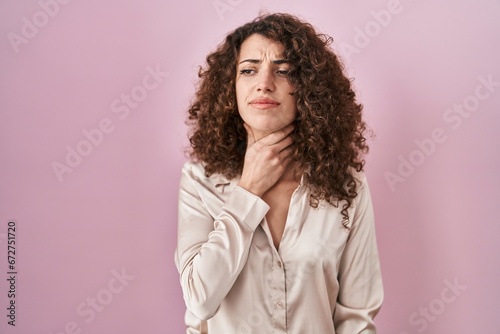 Hispanic woman with curly hair standing over pink background touching painful neck, sore throat for flu, clod and infection