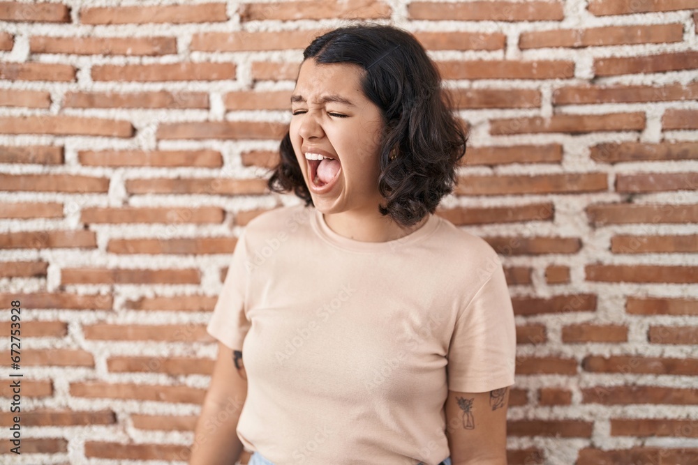 Young hispanic woman standing over bricks wall angry and mad screaming frustrated and furious, shouting with anger. rage and aggressive concept.