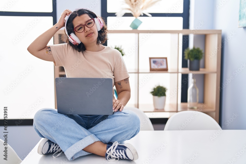 Young hispanic woman using laptop sitting on the table wearing headphones confuse and wonder about question. uncertain with doubt, thinking with hand on head. pensive concept.