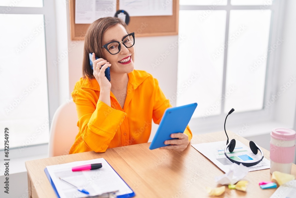 Middle age woman business worker talking on smartphone using touchpad at office Middle age woman business worker talking on smartphone using touchpad at office