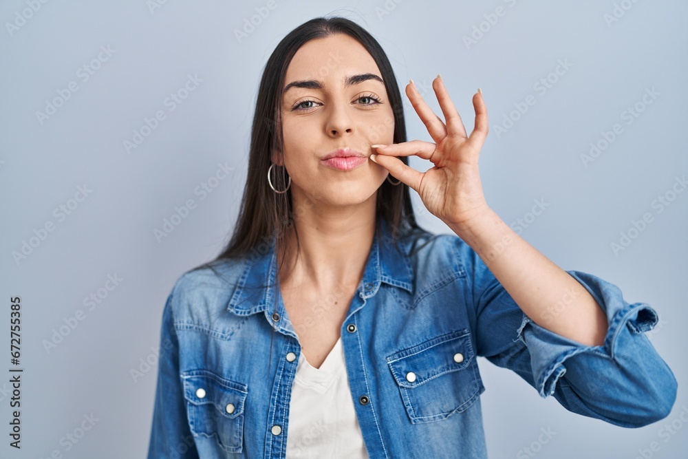 Hispanic woman standing over blue background mouth and lips shut as zip with fingers. secret and silent, taboo talking - obrazy, fototapety, plakaty 