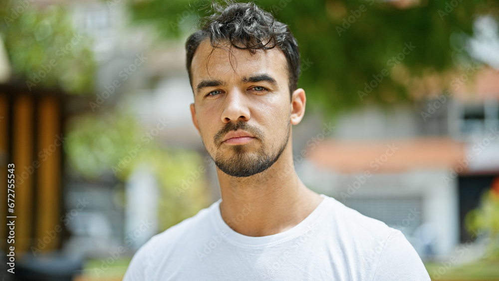 Young hispanic man standing with serious expression at park - obrazy, fototapety, plakaty 