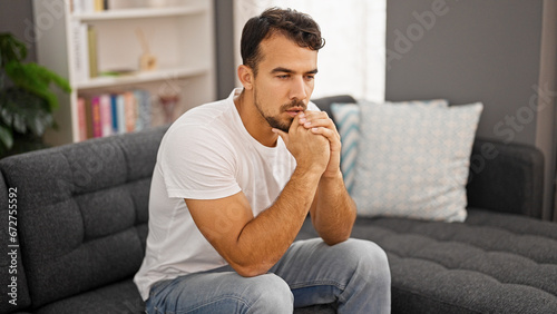Young hispanic man sitting on sofa with serious expression at home