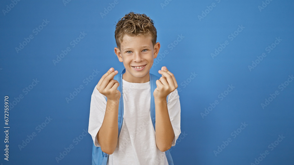 Adorable blond boy student, with a confident smile, joyfully doing a money gesture, standing isolated against a blue background with his smart academic bag Adorable blond boy student, with a confident smile, joyfully doing a money gesture, standing isolated against a blue background with his smart academic bag