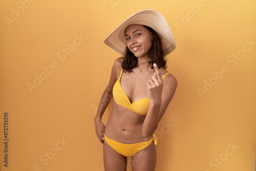 Young hispanic woman wearing bikini and summer hat beckoning come here gesture with hand inviting welcoming happy and smiling